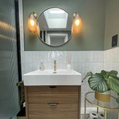 Monaco pearl brcik wall tiles fitted vertical straight stack in a green painted bathroom with wooden vanity, round brass mirror, feature lights, and plant on stand.
